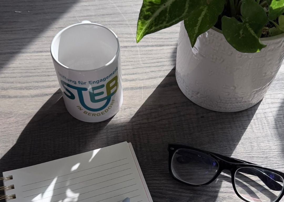 Abbildung von Schreibblock mit Stift, Brille und Tasse. Im Hintergrund eine Pflanze 